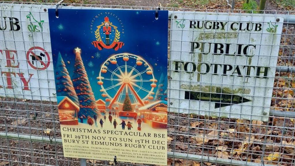 A close-up of a poster advertising the Christmas Spectacular Bury St Edmunds. It is attached to a wire fence, next to a sign giving directions to a footpath.  