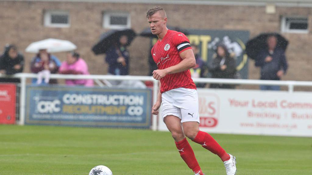 A man in a red football kit prepares to kick a ball on a football pitch with several people in a crowd watching on with umbrellas.