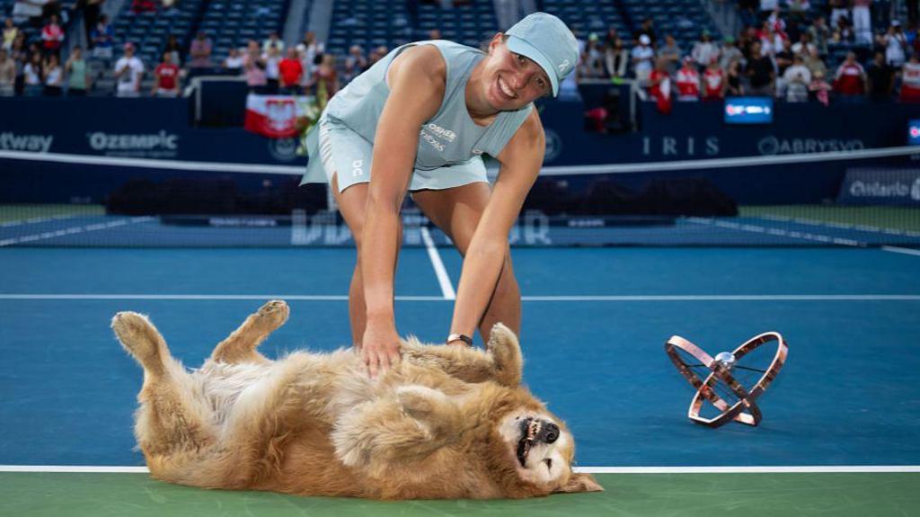 Iga Swiatek plays with a dog on court, as the Toronto trophy rests alongside them