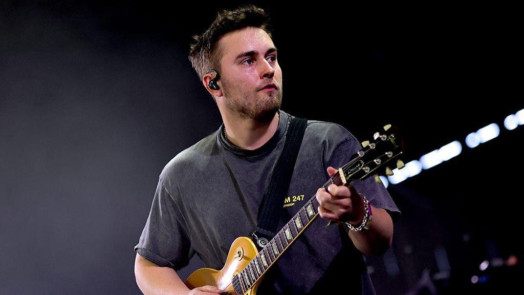 Sam Fender looks to his left as he plays a yellow electric guitar. He is wearing a dark t-shirt and has brown short hair and brown stubble. There is a dark black background.