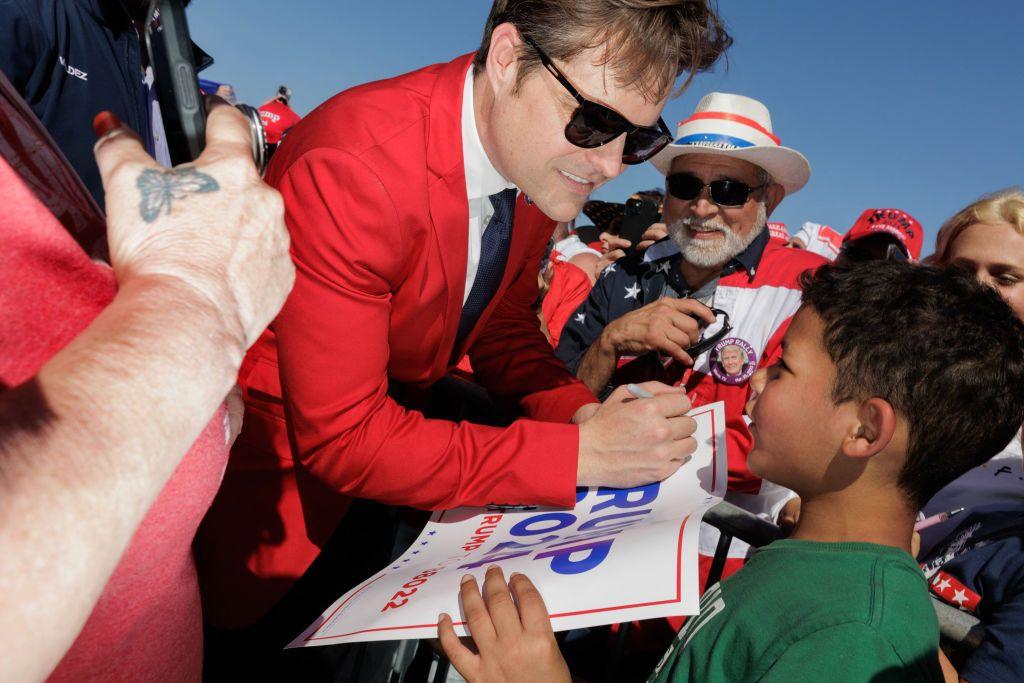 US Republican Representative Matt Gaetz signs posters and hats at a Trump rally in Texas in March. He is smiling at a child holding a sign with "Trump-Vance" on it. Gaetz is wearing sunglasses and a red blazer.