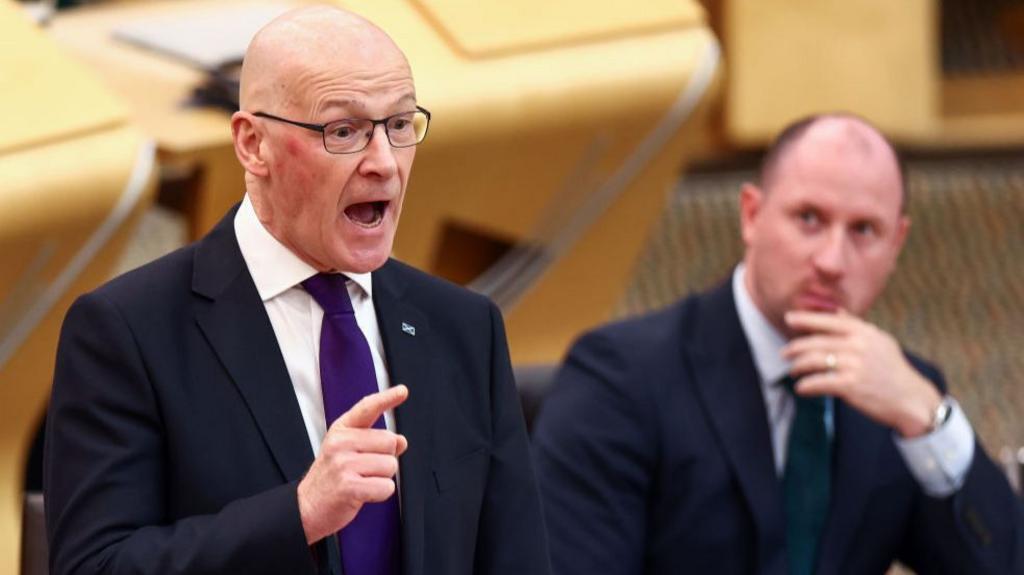 John Swinney has his hands out in front of him as he speaks in the Holyrood chamber. He is wearing glasses. He is wearing a dark suit, white shirt and purple tie. Health Secretary Neil Gray sits in the background.