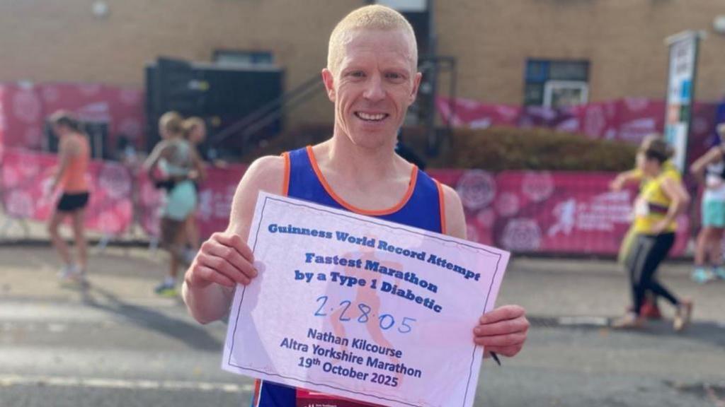 Image of Nathan in his running vest. He is holding up a sig which reads: GUINESS WORLD RECORD ATTEMPT FASTEST MARATHON BY A TYPE 1 DIABETIC: 2:28:05 NATHAN KILCOURSE ALTRA YORKSHIRE MARATHON 19TH OCTOBER 2025