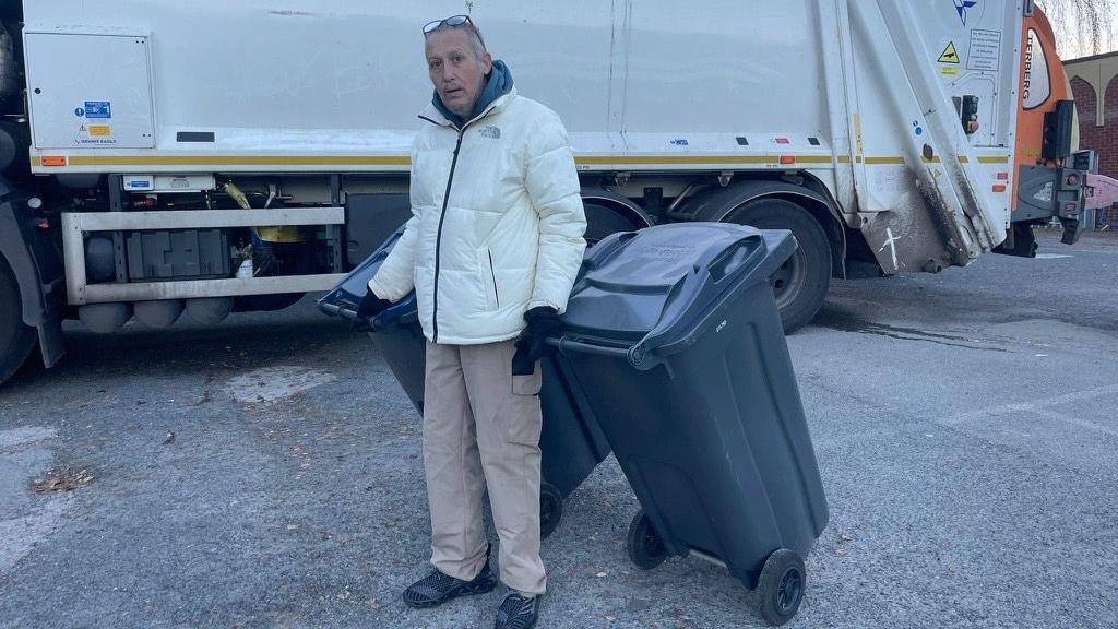 Resident Raymond McDonnell is pictured holding two dark blue recycling bins in front of a bin lorry. He has grey hair, glasses propped on the top of his head and is wearing a white North Face jacket, beige cargo trousers and black shoes.