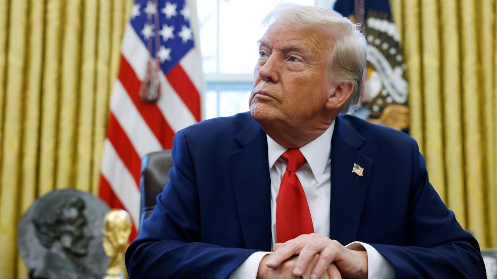 Donald Trump in the Oval Office. He is wearing a blue suit with a US flag pin. 