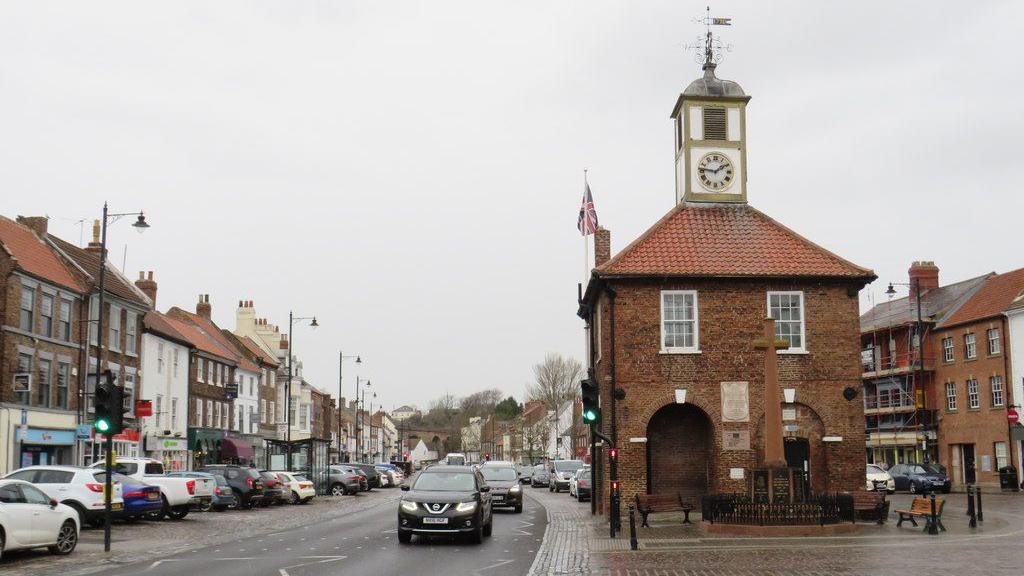 Yarm High Street, pictured on a cloudy and wet day. Cars drive along a small road next to the town hall building, which is a small hall with a clock tower. Shops are on either side of the street.