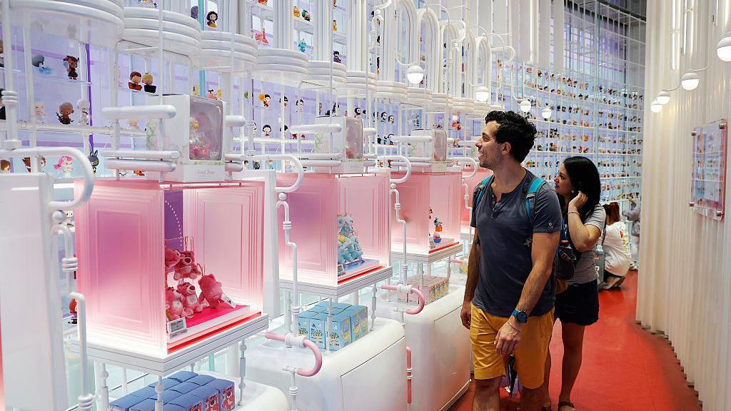 A man and a woman, look amazed, in front of a wall of Pop Mart dolls.