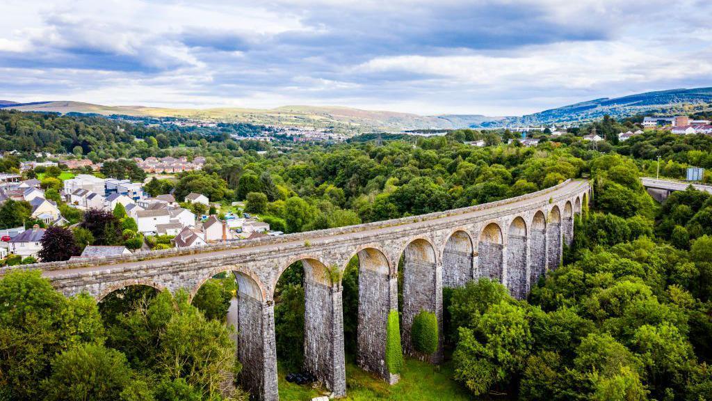 Cefn Viaduct with Merthyr Tydfil in the background - 13 large brick arches are in the foreground, surrounded by trees and rolling hills and housing in the background.
