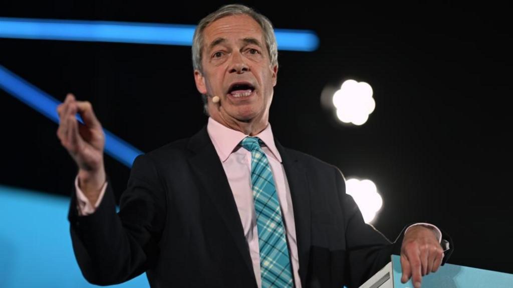 A close-up of Nigel Farage talking on stage, gesturing with one hand and resting the other on a lecturn. He is wearing a pale pink shirt, dark suit jacket and blue tie