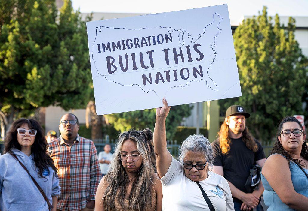 Protestors rally against an ICE deportation in Los Angeles, California. One protestor holds up a sign reading 'immigration built this nation' while bowing her head