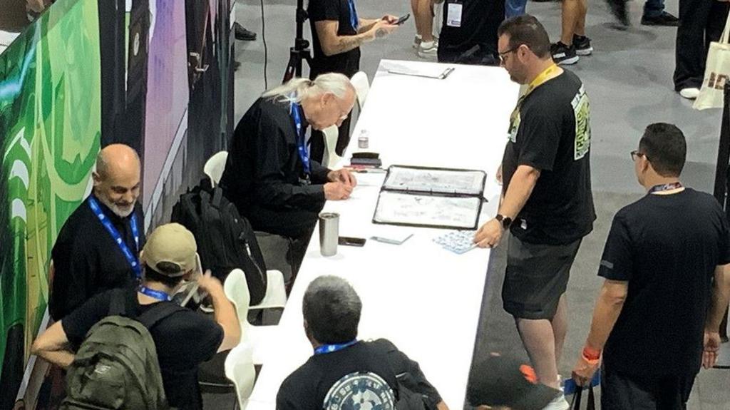 A man with long silver hair sits at a table signing art works for people