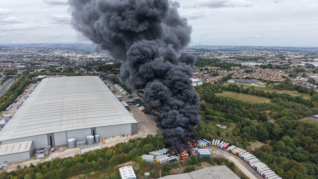 A wide angle shot of the HGVs on fire showing heavy smoke rising high into the air.