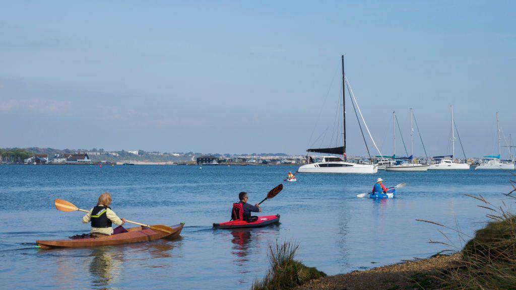 Group of people kayaking on the Christchurch Harbour, Dorset. You can see Mudford spit in the background with a large black building opposite Mudeford Quay. There are three people in kayaks wearing life jackets. There are several yachts moored in the still blue water of the harbour. 