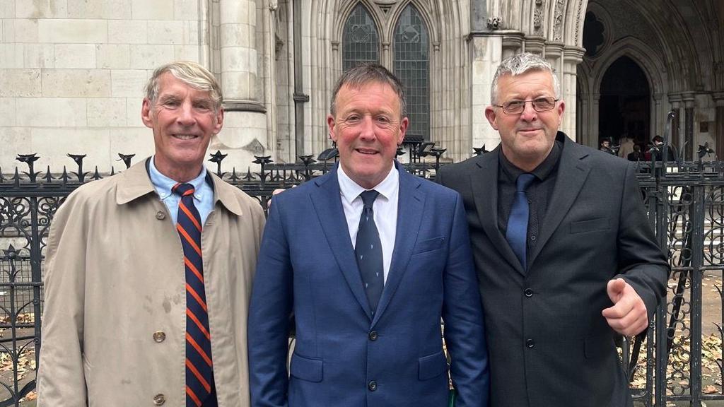 Left to right - Rob Sheridan in a blue shirt, blue and orange striped tie and a beige raincoat, Neil Stevens in a white shirt, navy blue tie and suit jacket, Tony Stevens in a black shirt with a navy blue tie and a grey suit jacket, all standing in front of the Royal Courts of Justice.