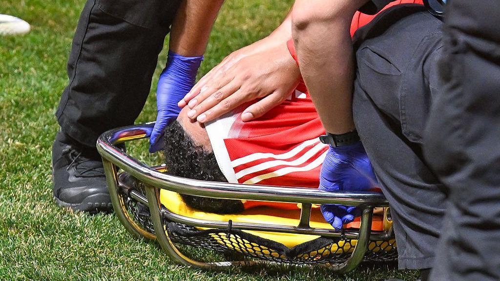 Musiala holds his shirt above his head while on a stretcher