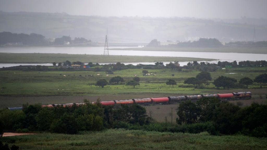 A train travelling through a rural area with a river and marshes seen in the background.