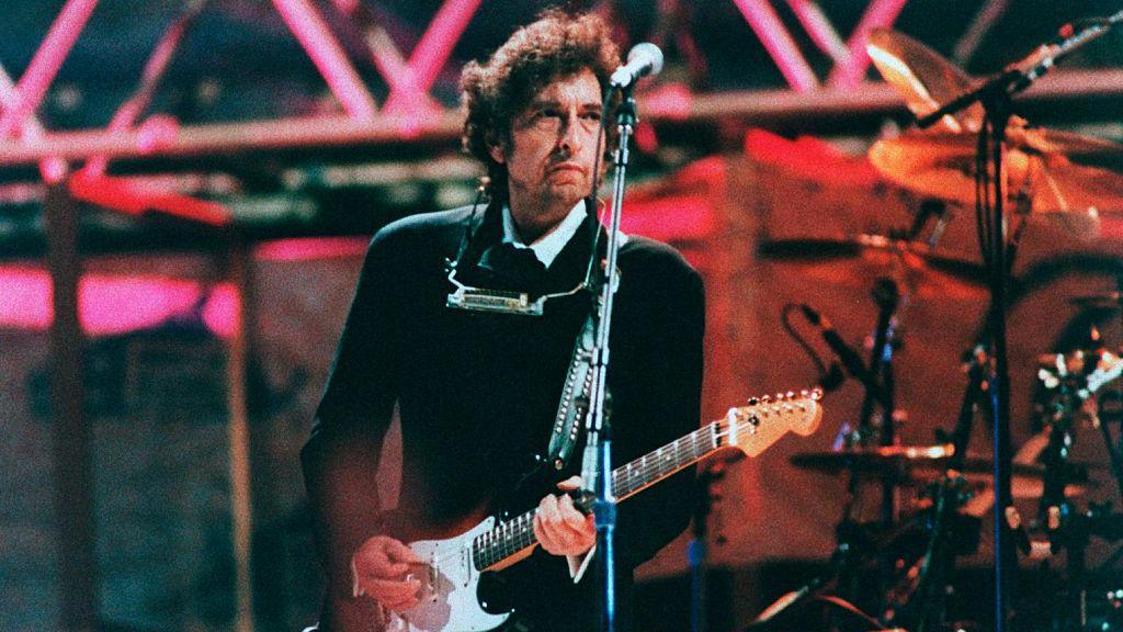 An old photo of Bob Dylan on stage in a black top with a white collar. He has brown curly hair. He is holding a guitar and has a harmonica around his neck. A microphone on a stand is in front of him, with drums and other microphones behind him and to his side