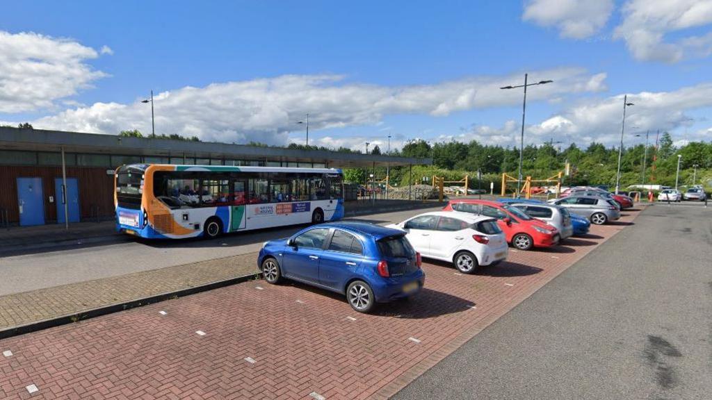 The large car park at Priory Park and Ride. Cars are parked in bays while a single-decker bus waits outside the single-storey bus station. A row of green trees can be seen in the background.