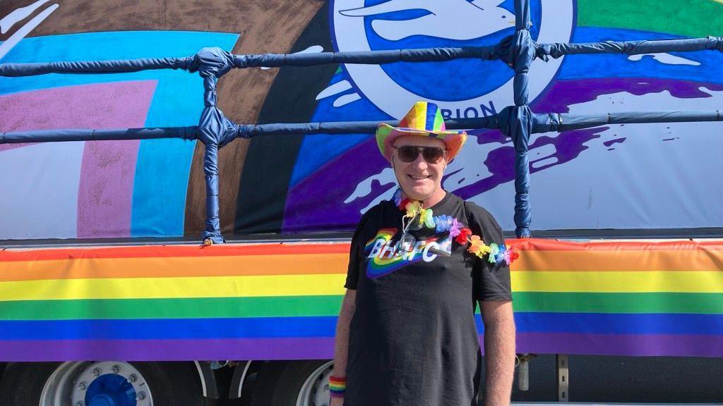 A man wearing a black shirt in front of a rainbow coloured float. His shirt has white writing reading "BHAFC". He is also wearing a rainbow hat.