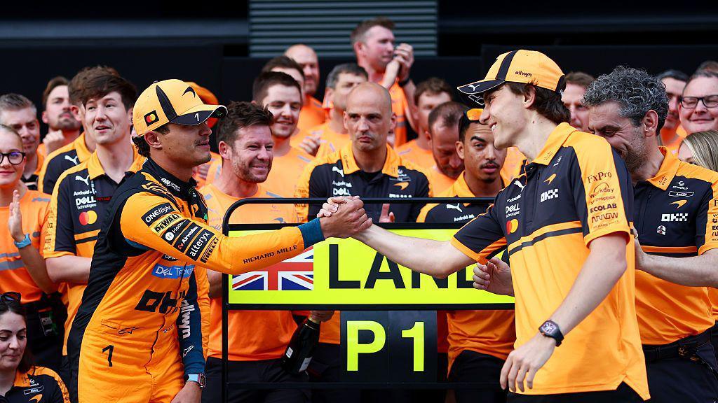 Land Norris and Oscar Piastri shaking hands in front of the McLaren staff waiting to take a team photograph