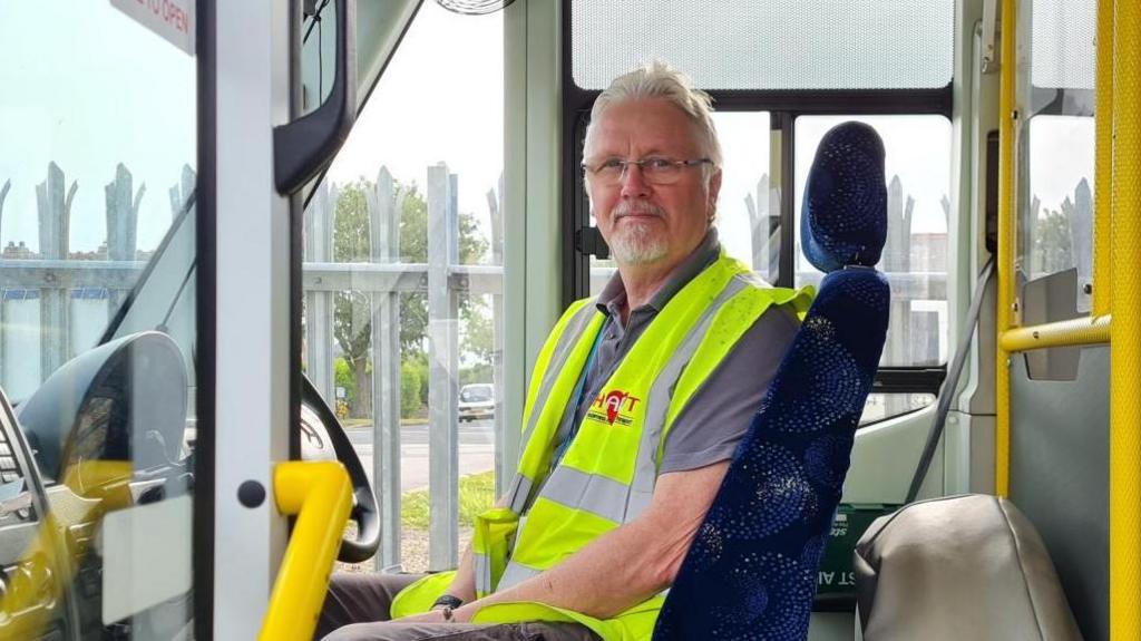 A man with a beard and grey hair seated behind the steering wheel of a minibus. He is wearing a yellow hi-vis vest. The vehicle is equipped with bright yellow grab rails.