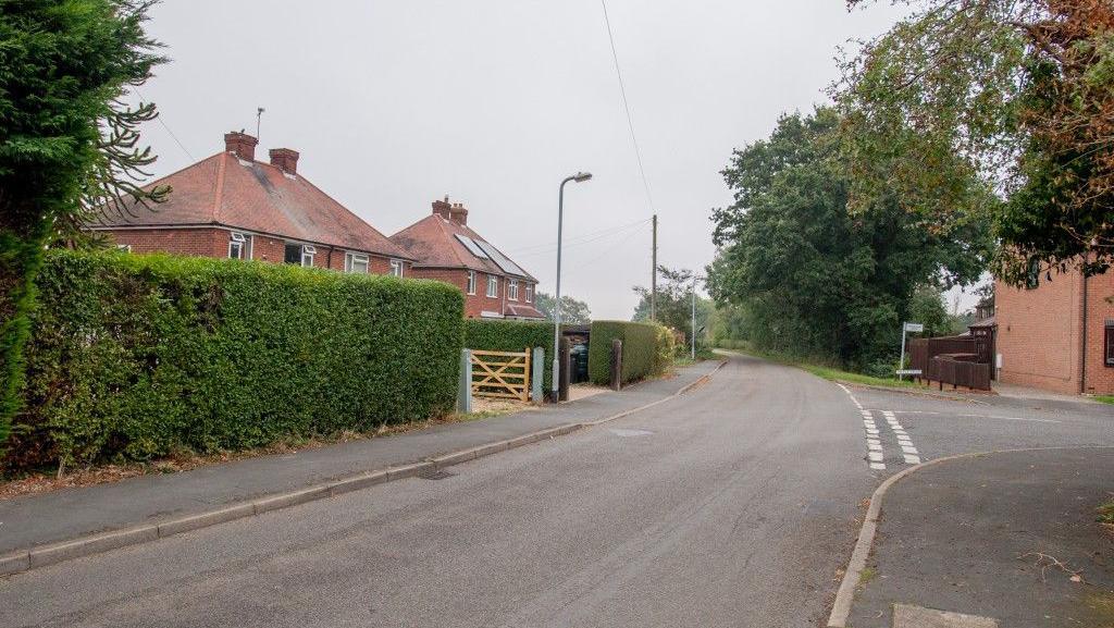 Shot of a quiet residential street with trees and houses. A wooden gate can be seen at the end of the driveway on one property and solar panels on the roof of another.