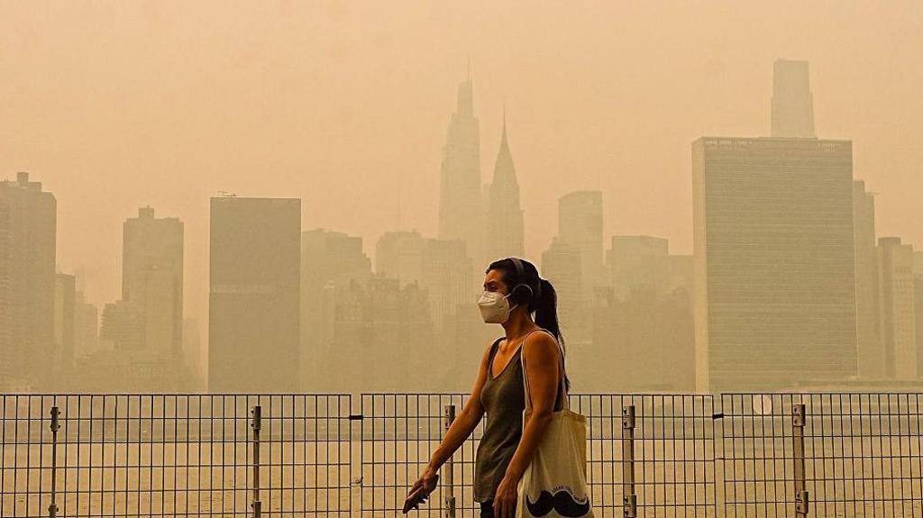 Woman with ponytail and mask walks in front of the New York City skyline, shrouded in an orange haze