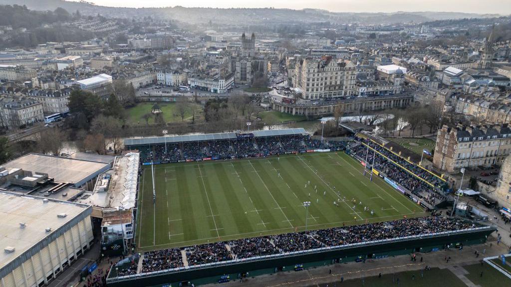 The Recreation Ground in Bath seen from above. There is a rugby game going on and the stands are filled with people. Behind the stadium are the hills of Bath.