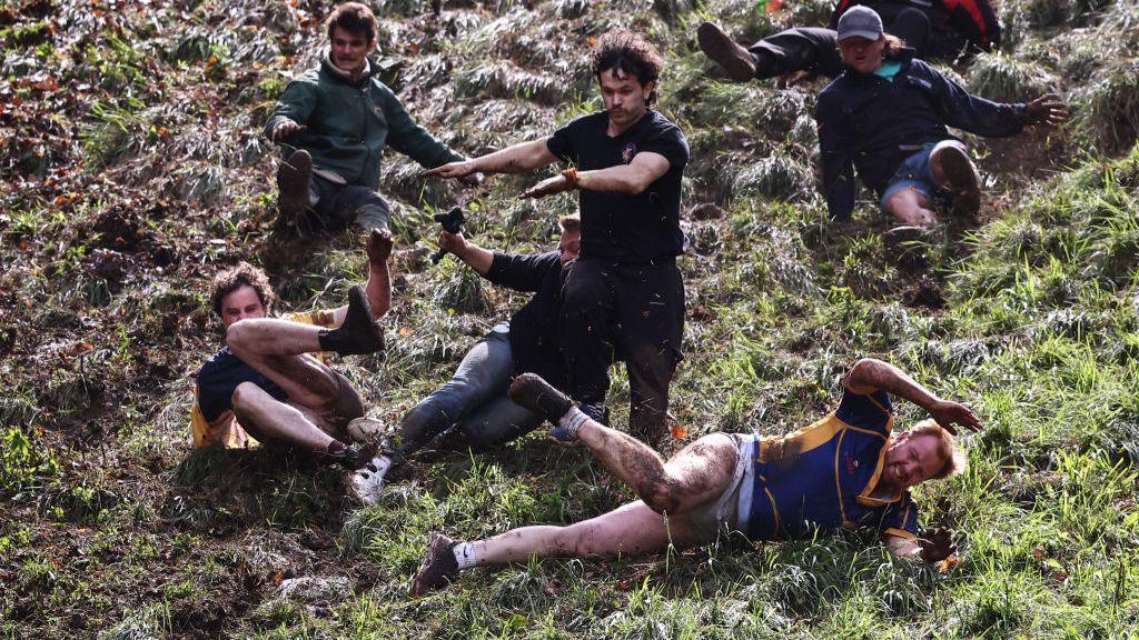 Seven men in various sport's clothing rolls down a grassy hill. Man at the forefront is wearing a jersey and shorts, his naked legs and arms covered in mud