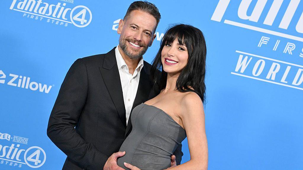 Gruffudd standing with Wallace at The Fantastic Four: First Steps World Premiere. Gruffudd is standing on the left, wearing a black suit. He has one hand on Wallace's bump and the other hand is behind her back. Wallace wears a bandeau grey dress and has black straight hair.