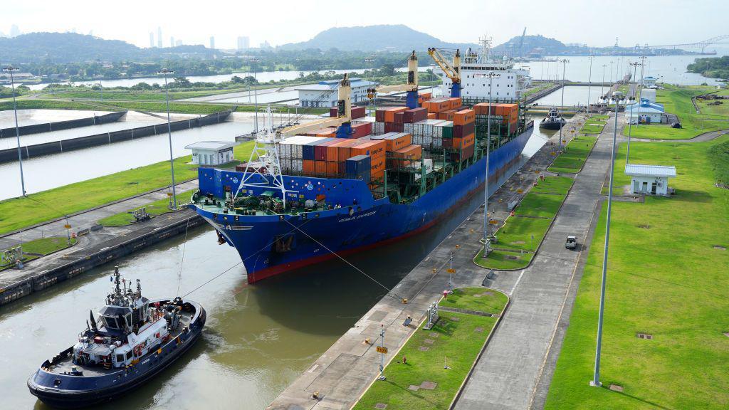 A large blue boat being pulled by a much smaller tugboat through a canal, with mountains in the distance