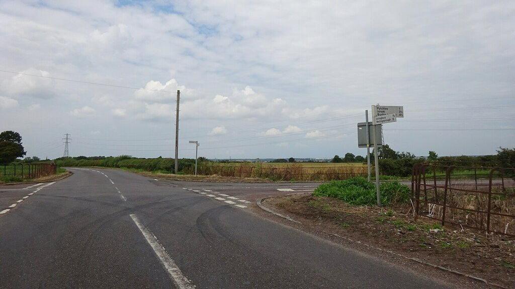 Four roads leading into a crossroads junction. There are green fields on either side of each road. The sky is cloudy, and there is a direction sign in the foreground.