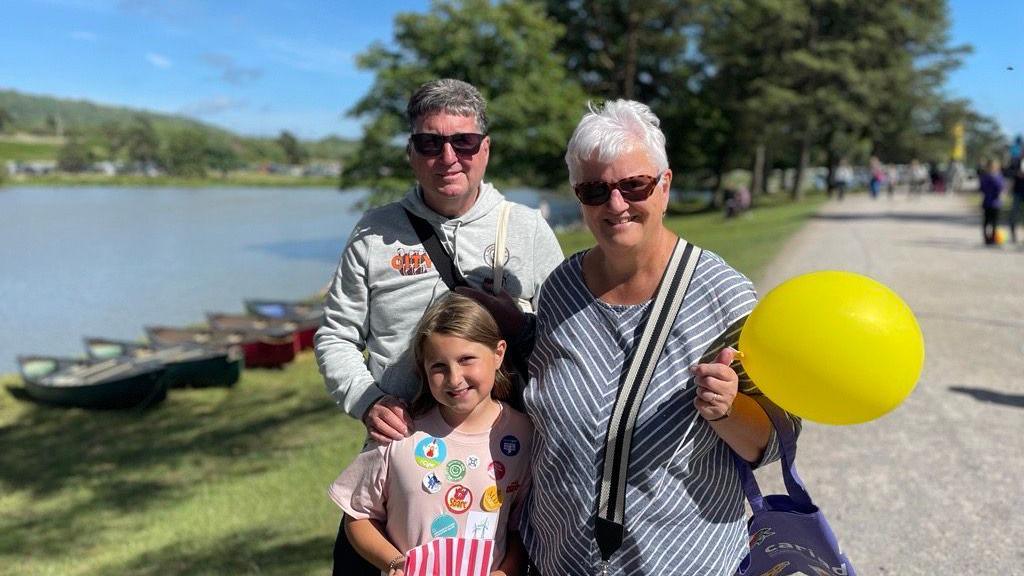 Dilys, Arwyn and Mai stand in front of the lake with trees behind them and blue skies beyond. Both grandparents are wearing sunglasses, Arwyn has his hand on Mai's shoulder. Dilys is holding a bright yellow balloon.  Arwyn has a grey hoody on and dark grey hair, Dilys has white hair and has a blue and white striped top with a bag strap on her shoulder which is black and white. Mai is wearing a pink t-shirt with loads of badges on it.