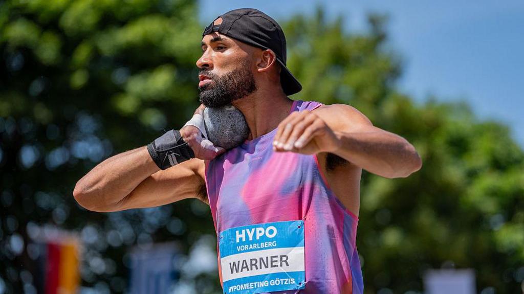 Decathlete Damian Warner holds a grey shot-putt under his chin with his right hand as he prepares to throw, wearing black back-to-front baseball cap, tie-dye sleeveless vest and competitor's label on chest bearing his surname