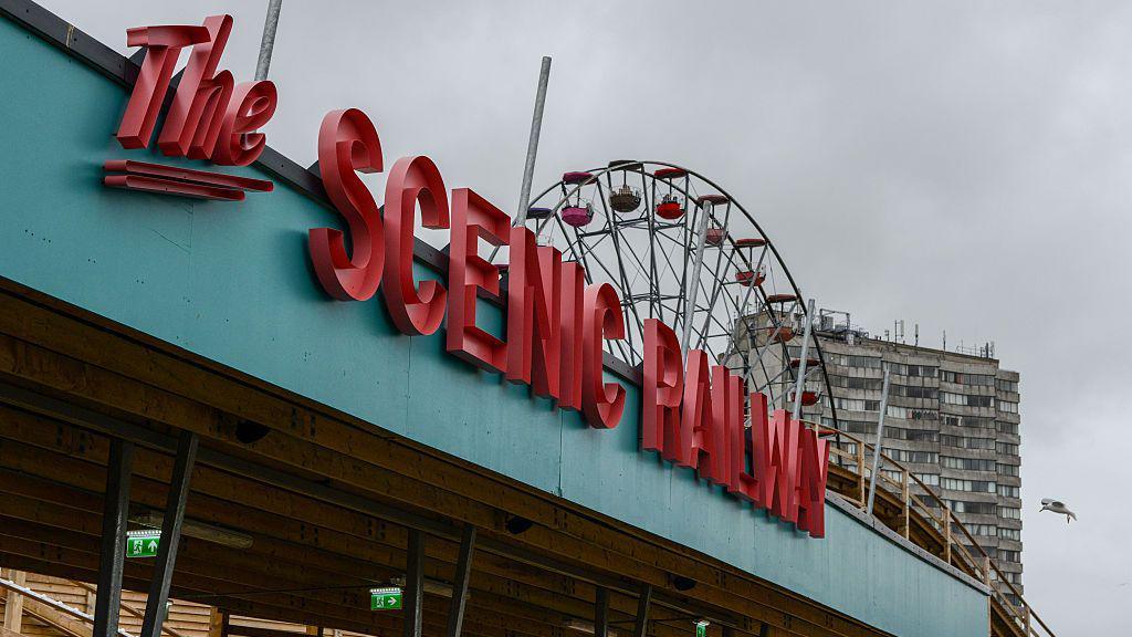 Shkronja të mëdha të kuqe që shkruajnë "Hekurudha piktoreske" në një çati blu. Në sfond mund të shihen pjesë të një rollercoaster prej druri, një rrotë panoramike dhe një kullë e madhe.