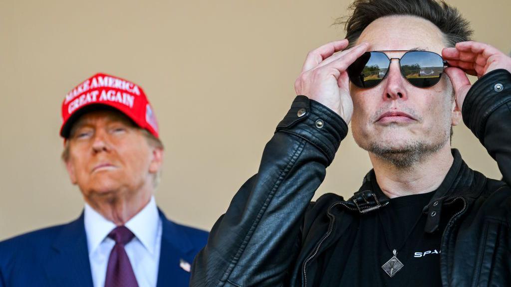 Mr Trump stands beside Mr Musk. Trump is wearing a red Make America Great Again cap and a sharp blue suit. Musk is wearing a leather jacket and aviator sunglasses.