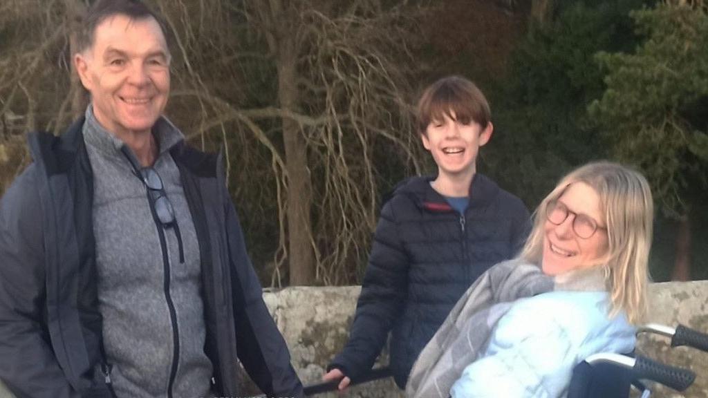 Janine with her husband and 11- year-old son. Janine has shoulder length blonde hair and is in a wheelchair, with her husband and son standing alongside her. They are all smiling. 