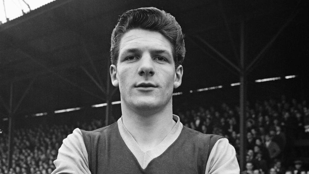 John Lyall when he was playing for West Ham in a black and white photo. He stands on a football pitch in front of a crowd of people who are in the stands. He has his arms crossed in front of his chest.