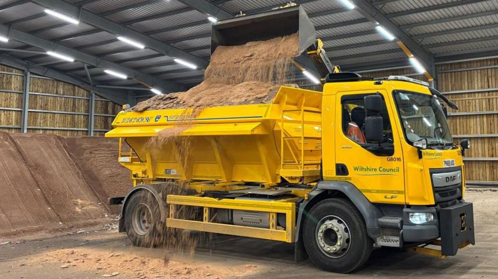 A large yellow gritter truck with a bed overflowing full of sand, salt and grit. It is parked inside a large metal warehouse with white overhead lighting. Behind the truck there is a pile of grit.