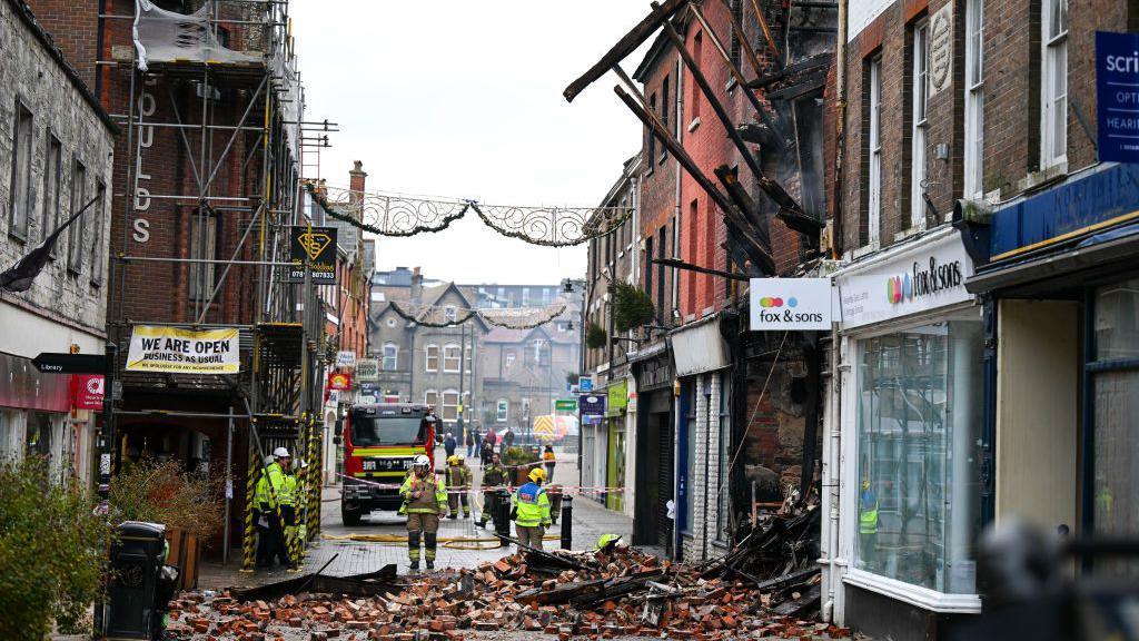 A general view down South Street, with firefighters and a fire engine standing in front of a pile of bricks that have fallen with the building.