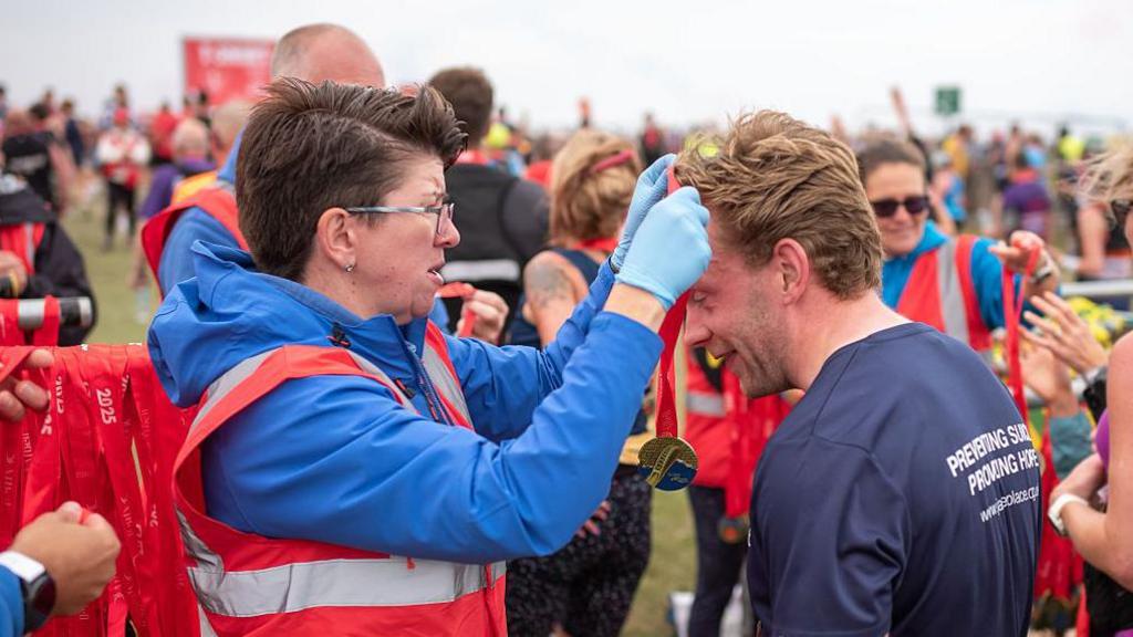 A male runner is presented with a medal at the finishing line. He has fair hair and is wearing a blue t-shirt. The woman hanging the medal around his neck has short dark hair, glasses and a light blue jacket.