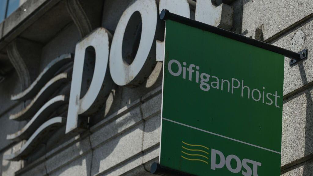 A green sign which says, in white writing, 'Oifig an Phoist'. Below that, there is a white line on the sign, and below that is written 'post', with four horizontal yellow lines to the left of it. The sign is in front of a brick building, which has a large stone sign saying 'post' on the front. 