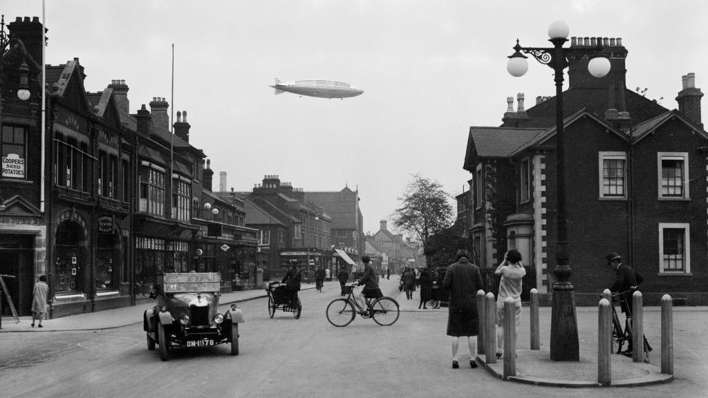 The R101 airship flying over the town of Bedford on its first flight. It is a black-and-white image of a busy street, with shops, cars. People are walking in the street and riding bicycles. People have stopped to look up at the airship.