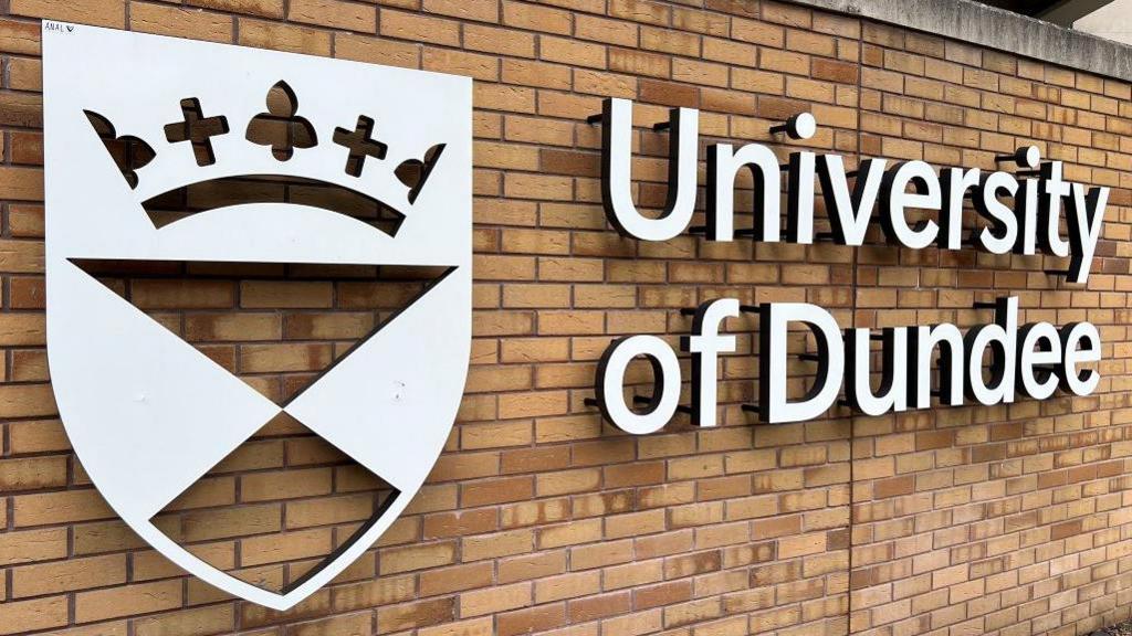 A white sign on a brown brick wall with a crest and the words "University of Dundee"