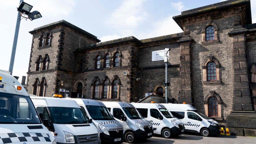 A general view of a Serco vehicle at Wandsworth prison in London. It is an imposing building with a gatehouse and what looks like a portcullis. A row of prison vans are by the entrance