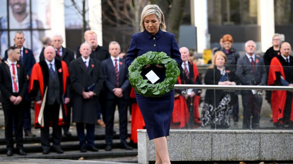 Michelle O'Neill in navy skirt suit and black heels, laying a green laurel wreath at remembrance event in Belfast City Hall, out of focus crowd in background watch on in dark clothes