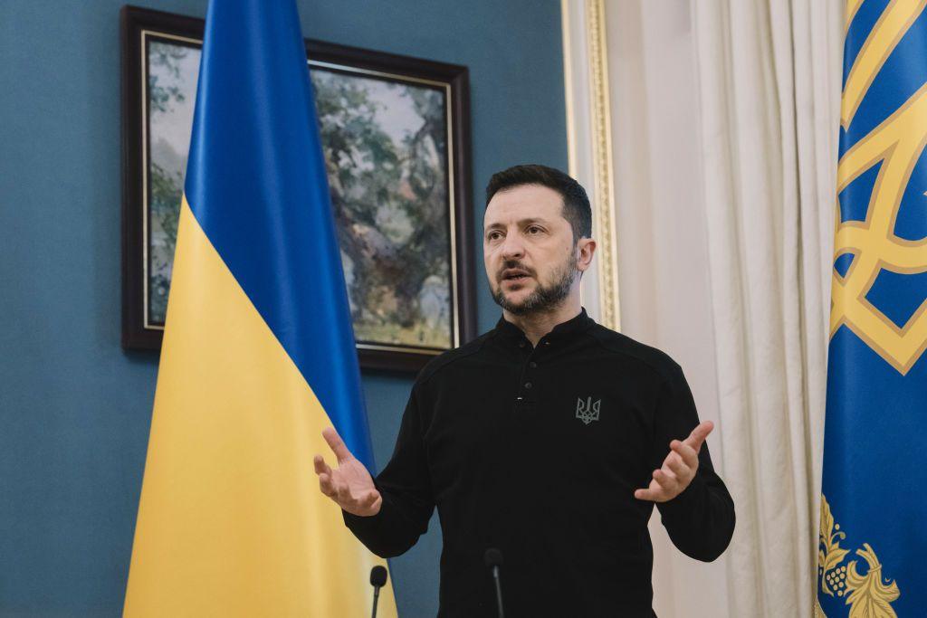 President Zelensky, hands raised, mid-speech, pictured in front of Ukraine flag