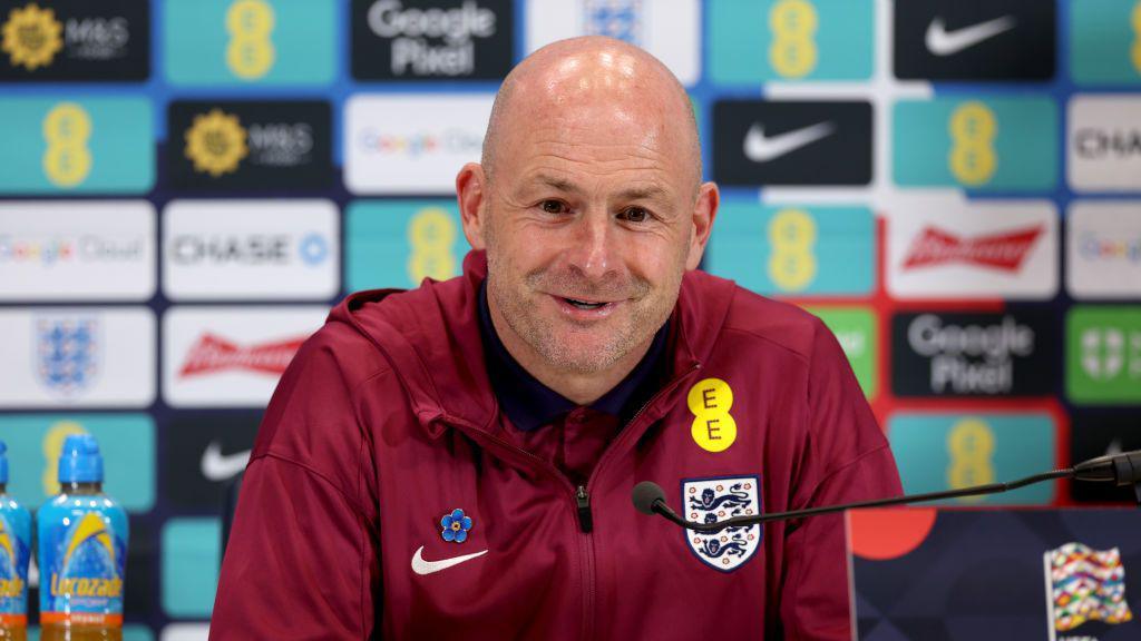 Lee Carsley in a maroon red England jacket, sitting at a table with a microphone in front of him. He is smiling. He is bald and has a blue flower pinned on his jacket