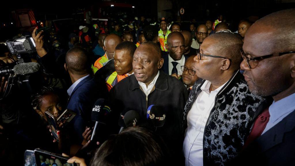South African President Cyril Ramaphosa speaks to journalists at the scene of the building fire. It is dark, but he is lit by camera lights. The president is surrounded by crowds of security guards and other politicians.