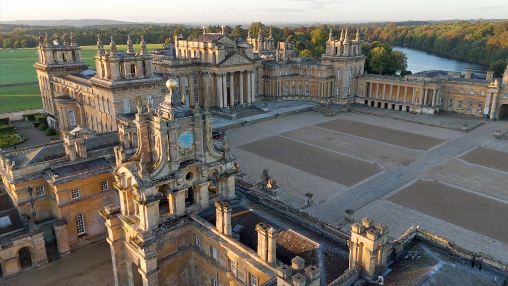 An aerial image of Blenheim Palace - showing a large courtyard surrounded by baroque buildings in yellow stone, with ornate turrets and chimneys. Woods, fields and a river can be seen surrounding it.